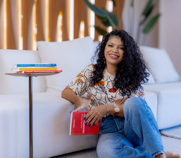Josiane sentada no chão do consultório, apoiada no sofá, com um livro de psicoterapia nas mãos