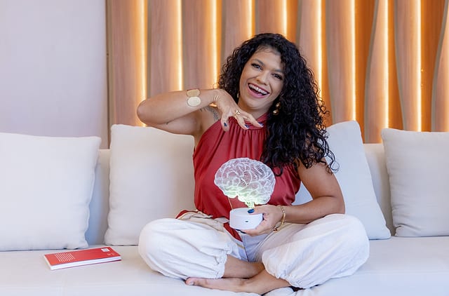 Josiane sentada na cama, sorrindo, com uma almofada em formato de cérebro no colo